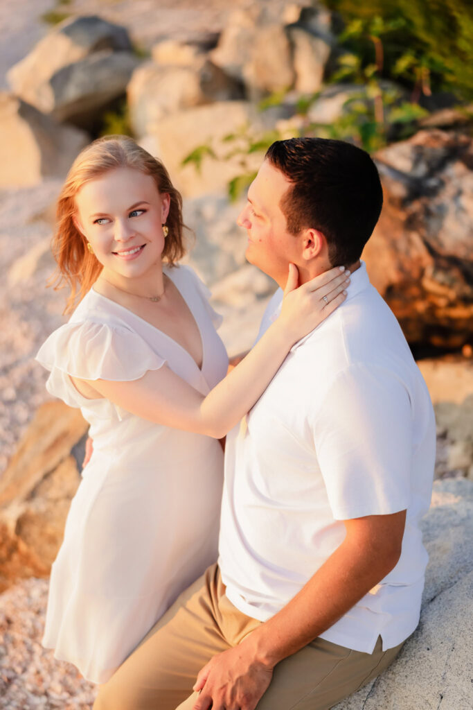 Bride-to-be sitting on a rock, smiling lovingly at her fiancé at Greenwich Point Park.