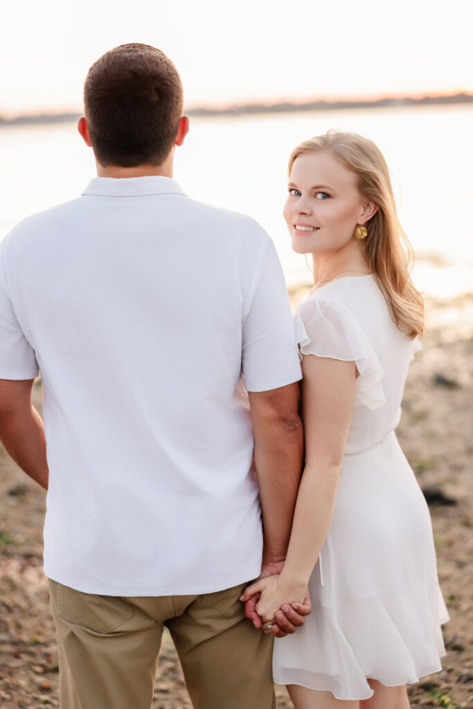 Bride-to-be walking hand in hand with her fiancé along the beach, looking over her shoulder at Greenwich Point Park.