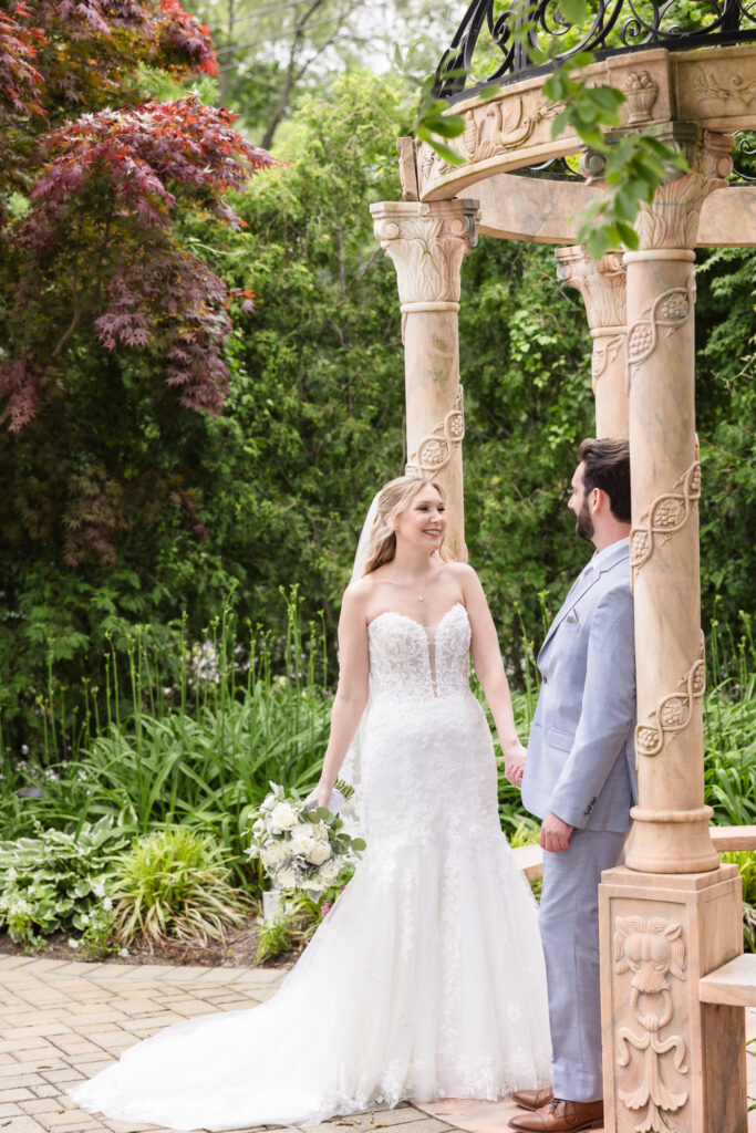 Bride and groom standing face to face under a floral arch, framed by lush greenery and soft daylight.