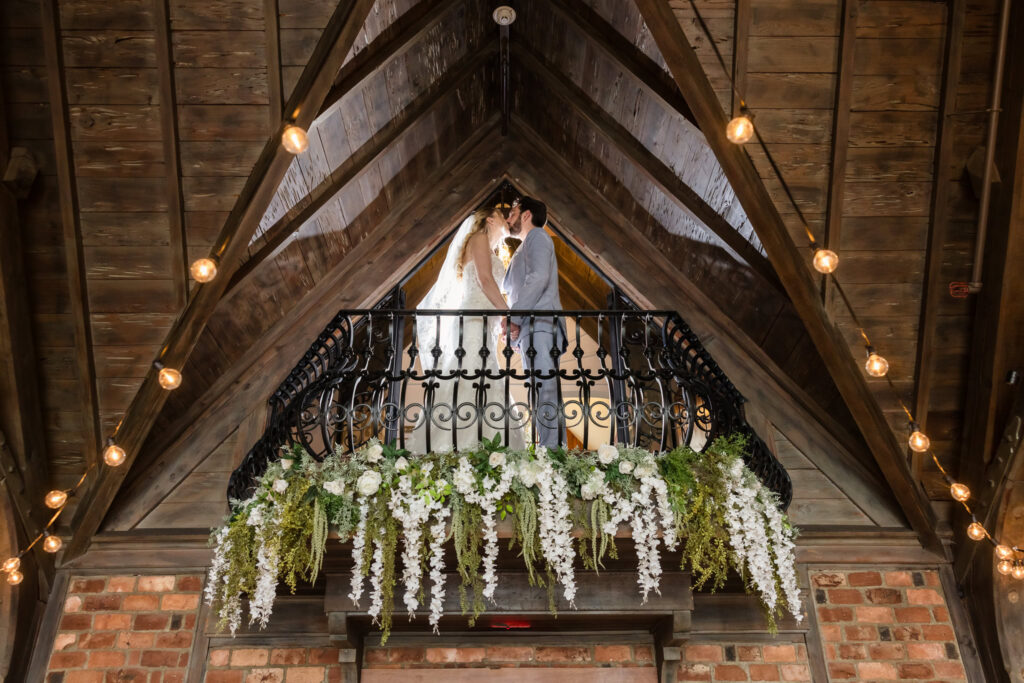 Bride and groom standing together on a balcony adorned with hanging florals inside the rustic Riviera Waterfront Mansion venue.