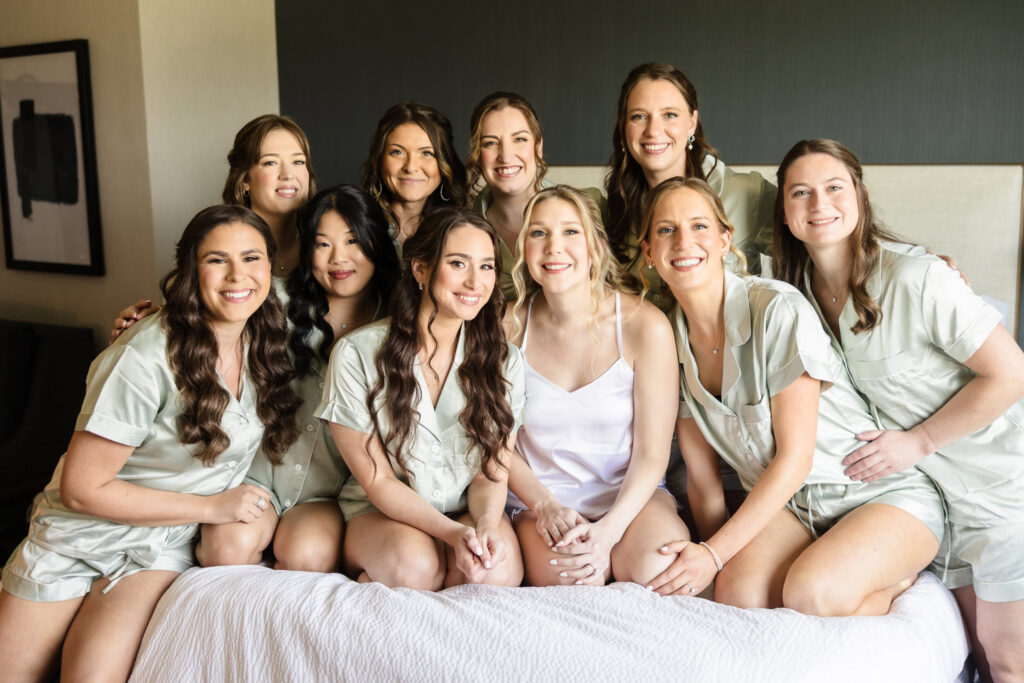 Bride sitting on a hotel bed surrounded by her bridesmaids in matching robes, sharing laughter and excitement before the ceremony.