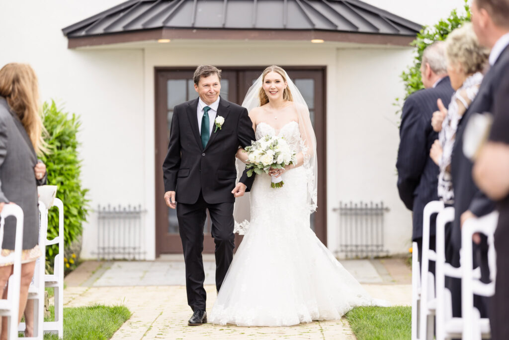 Bride walking down the outdoor aisle with her father, smiling as guests watch lovingly.