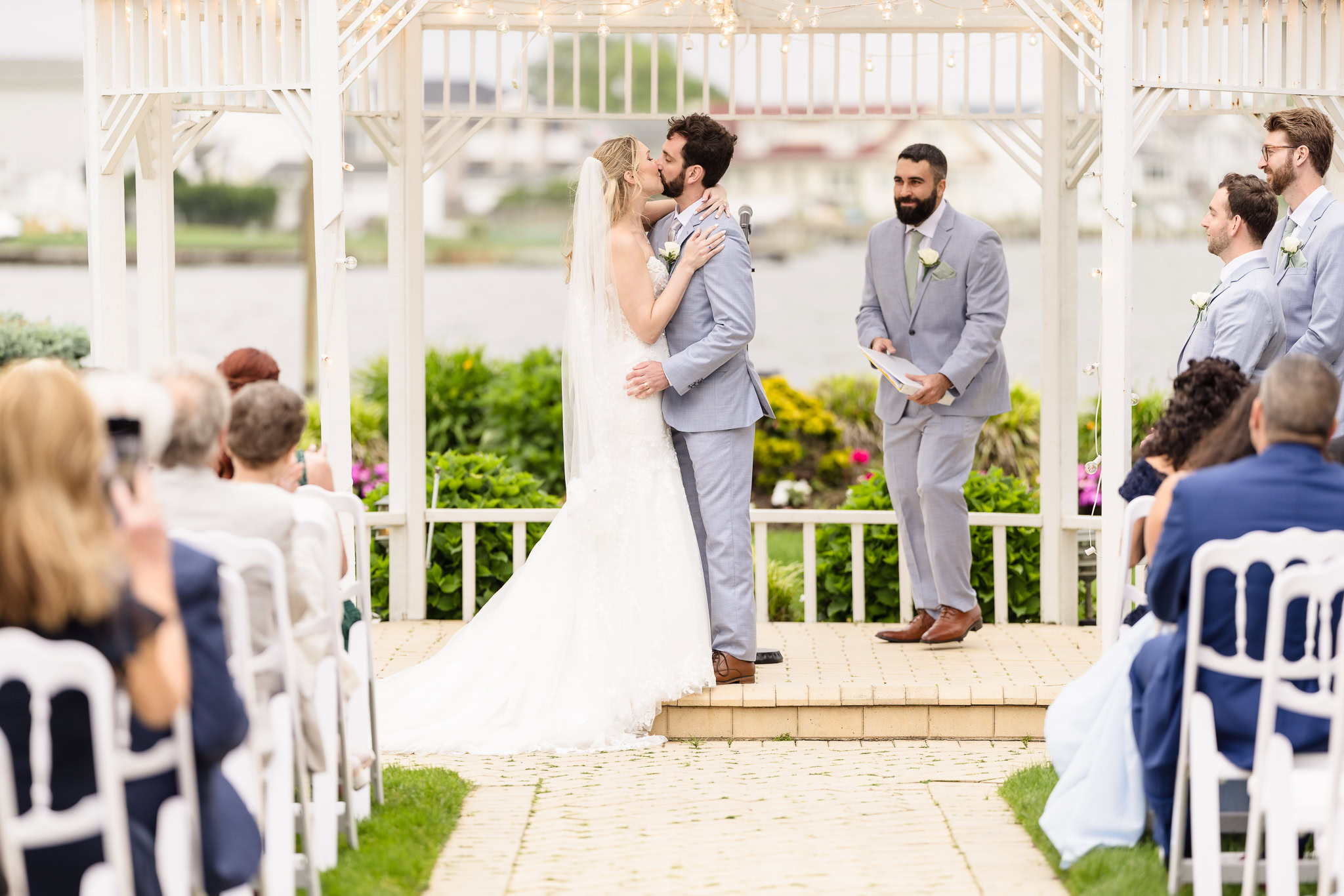 Wide shot of the outdoor wedding ceremony, guests seated as the couple stands with their officiant beneath a bright sky.