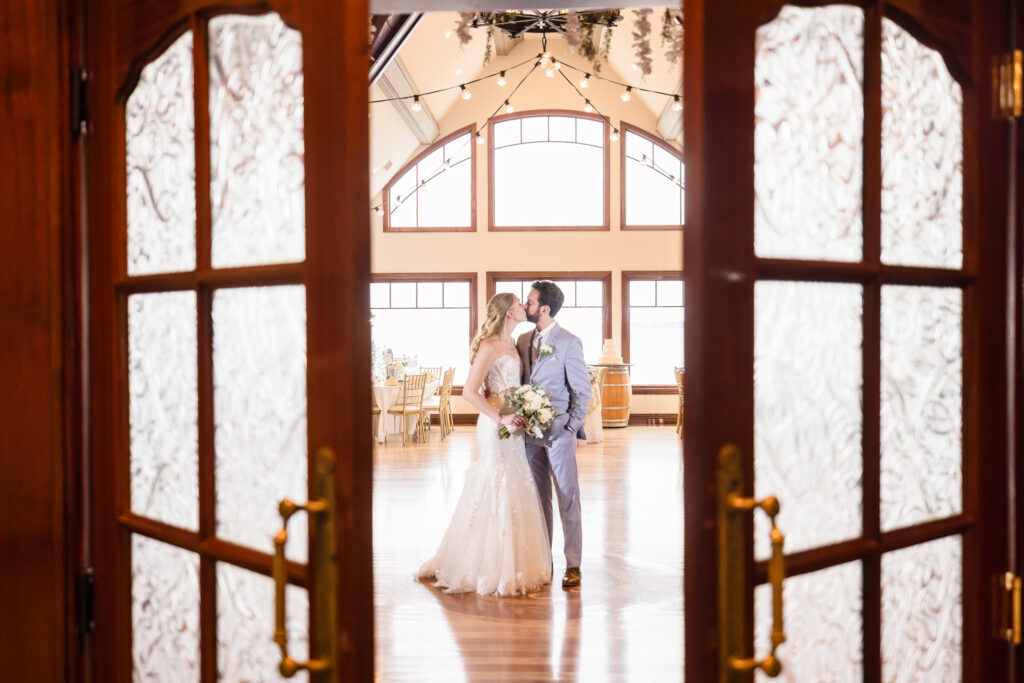 Bride and groom sharing their first dance inside Riviera Waterfront Mansion, framed through elegant glass doors.