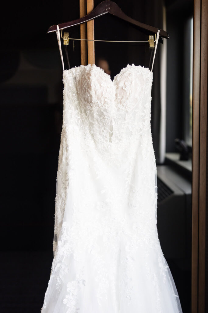 Lace wedding gown hanging on a wooden hanger by a dark doorway, softly lit to highlight its elegant detailing.