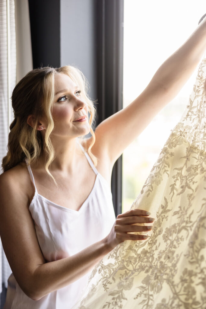 Bride holding her lace wedding dress up to the window, smiling as sunlight filters through the delicate fabric.