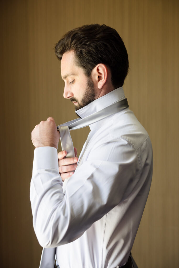 Groom adjusting his cufflinks while getting dressed in a white shirt, softly lit in a calm preparation moment.