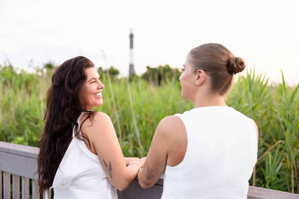 Couple smiling at each other, surrounded by tall green beach grass with the distant Robert Moses lighthouse visible behind them.
