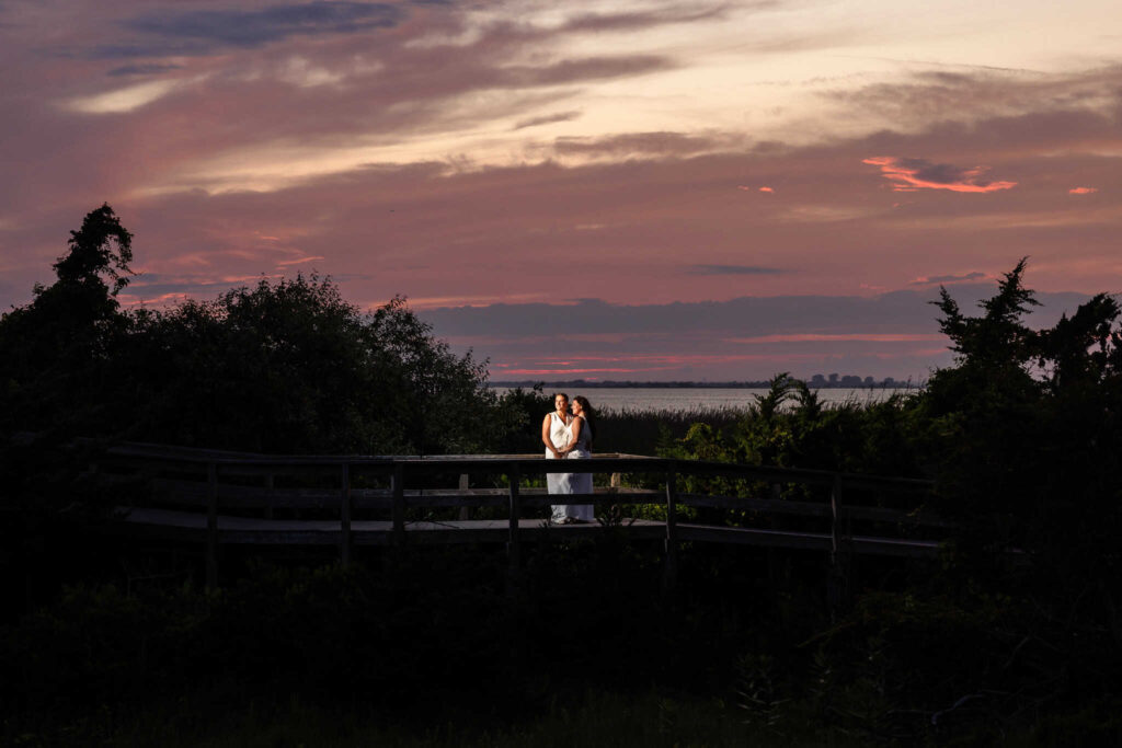 Couple holding hands and leaning close on a wooden walkway, framed by deep pink and purple hues of a summer sunset sky.