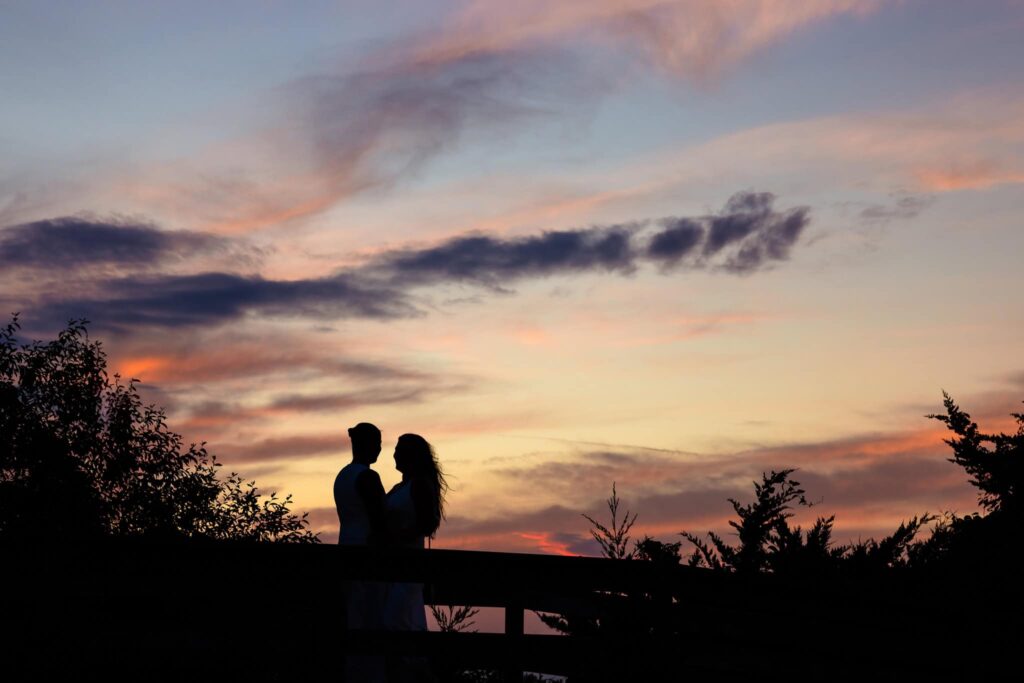 Couple standing close together, silhouetted against a dramatic sunset sky with streaks of gold and violet at Robert Moses State Park.