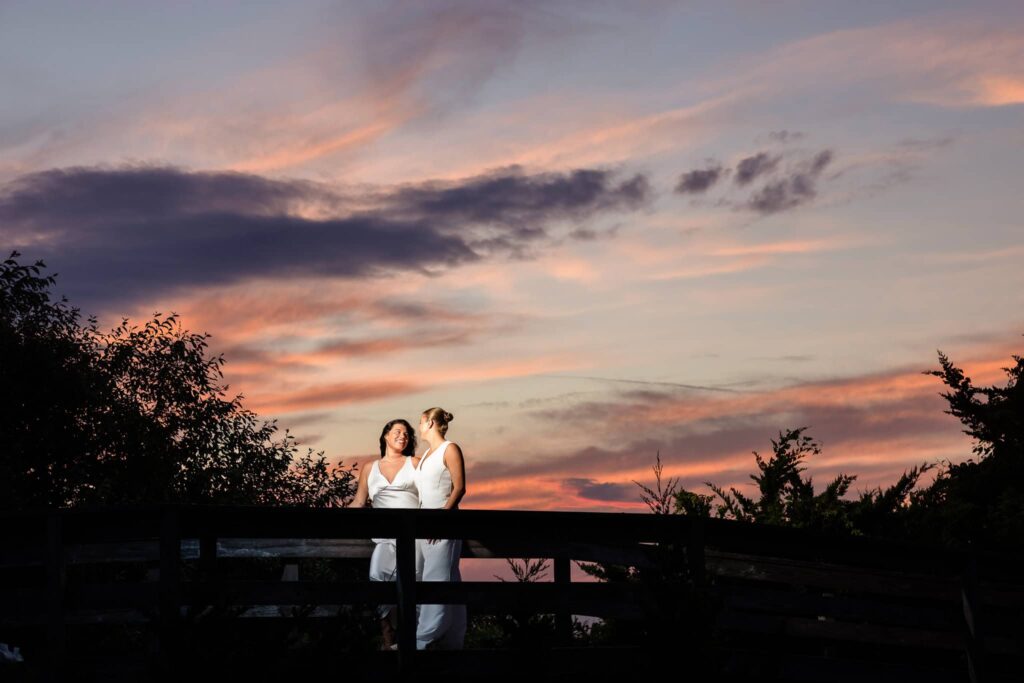 Couple embracing on a wooden boardwalk with vibrant orange and purple clouds stretching across the evening sky.