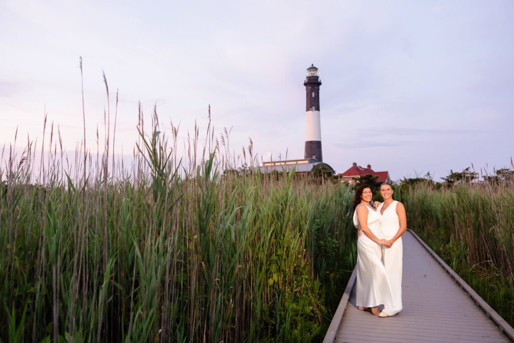 Couple walking hand in hand through tall dune grass, the Robert Moses lighthouse standing in the distance beneath a fading pastel sky.
