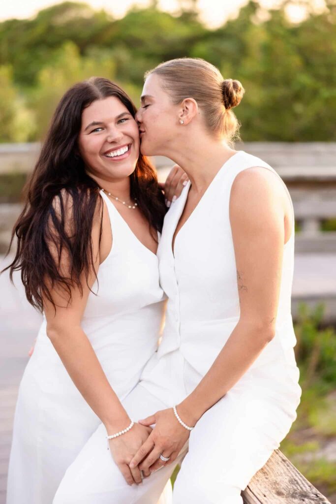 Couple sitting close together on a wooden ledge by the dunes, laughing and leaning into each other in a quiet, intimate moment.