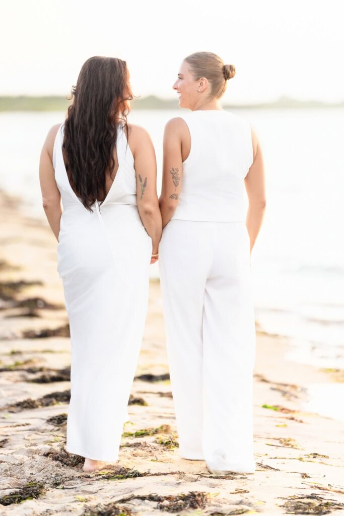 Couple standing barefoot on the sandy shoreline, holding hands and gazing at the water as gentle waves meet the beach at dusk.