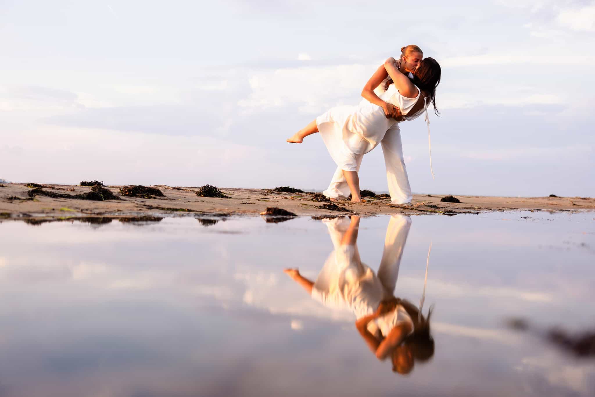 Couple sharing a joyful dip kiss by the water’s edge, their reflection mirrored in a still tide pool at Robert Moses State Park.