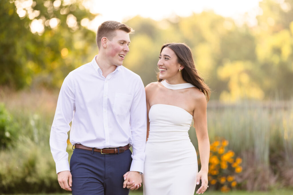 Couple holding hands and smiling at each other while walking through a garden at sunset.