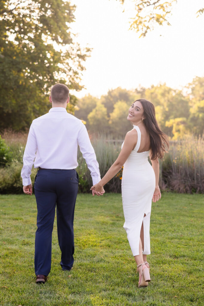 Woman looking back and smiling while walking hand in hand with her fiancé through a garden.