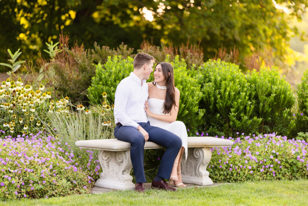 Couple sitting on a stone bench surrounded by garden flowers during their engagement session.
