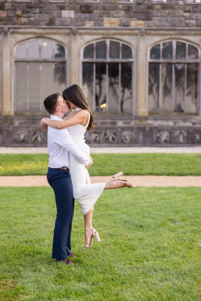 Fiancé lifting his bride-to-be for a joyful kiss in front of a castle-like mansion facade.
