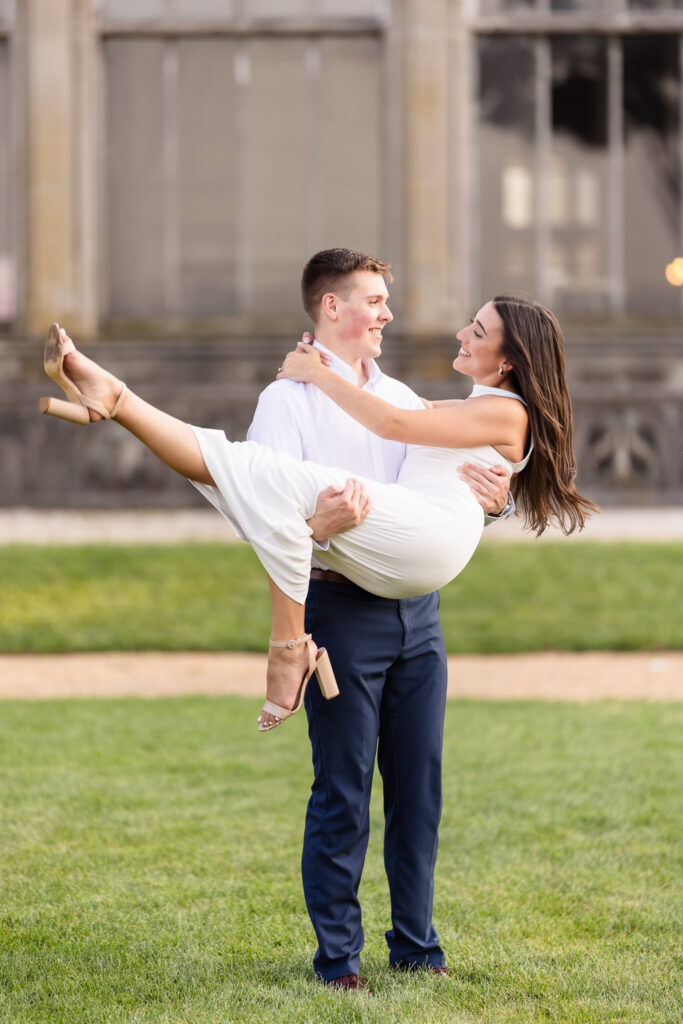 Groom-to-be carrying his fiancée in his arms, both smiling joyfully in front of the historic mansion.
