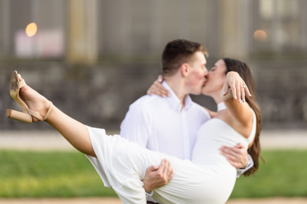 Couple kissing while she is held in his arms, her hand extended to show off the engagement ring.