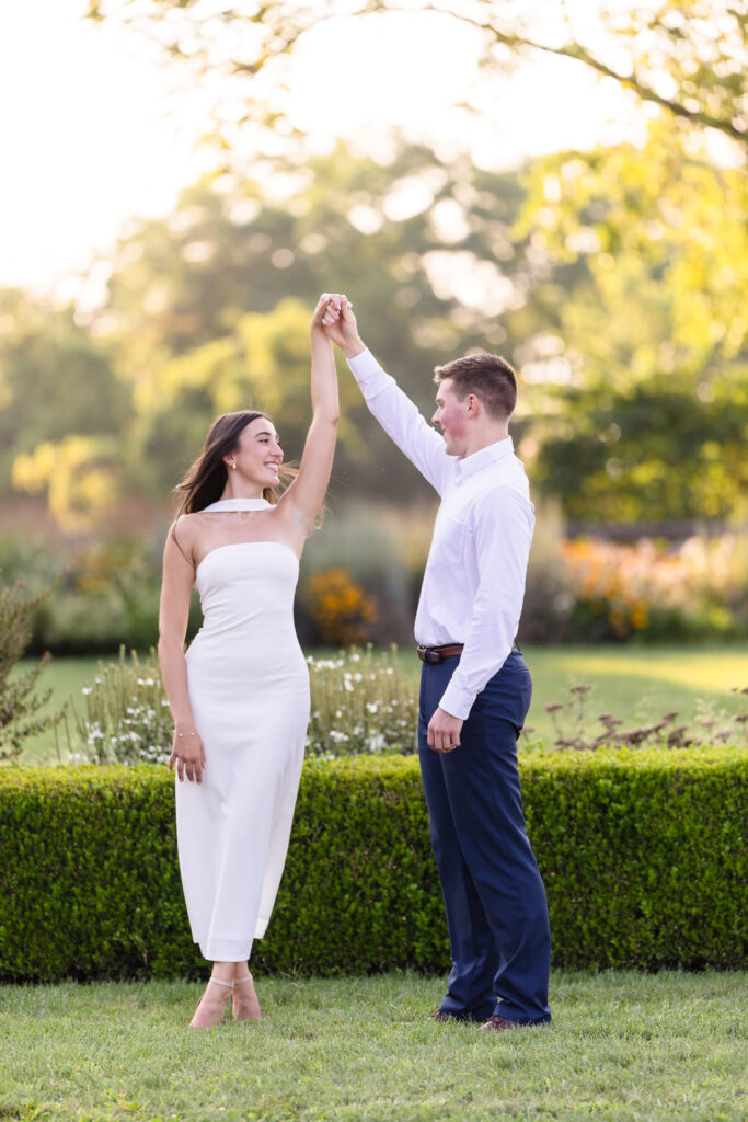 Couple holding hands as he twirls her gently, surrounded by greenery and golden evening light.