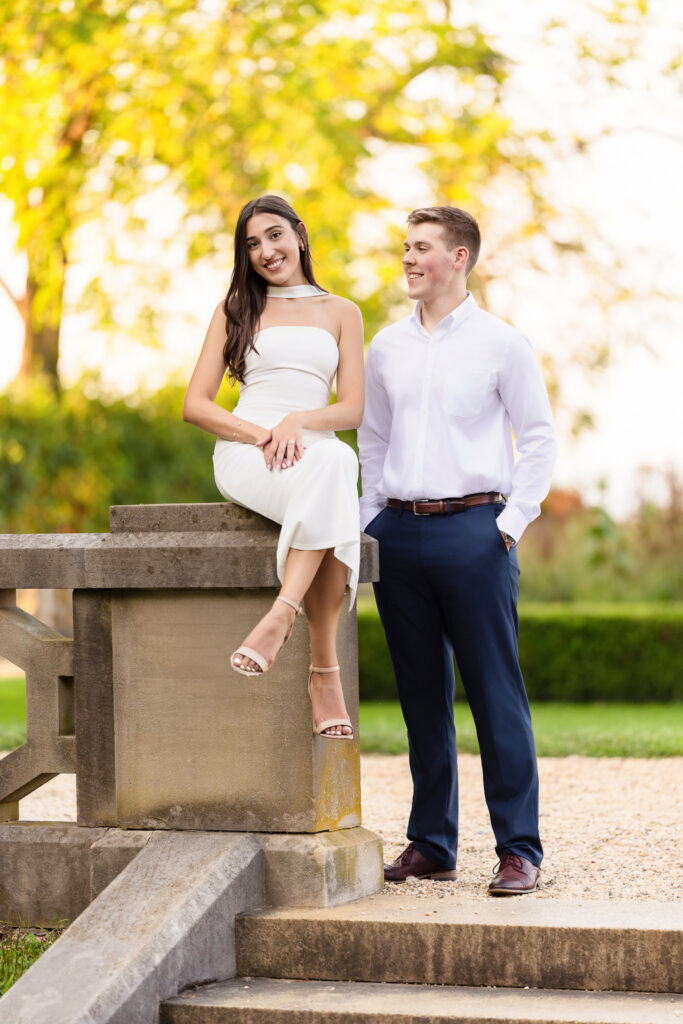 Bride-to-be sitting gracefully on a stone ledge while her fiancé stands beside her, both smiling.