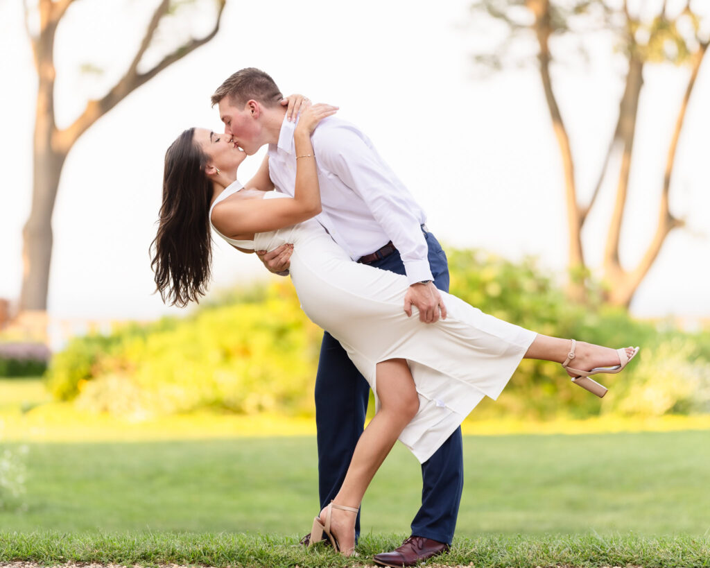 Groom-to-be dipping his fiancée for a romantic kiss on the lawn surrounded by lush greenery.