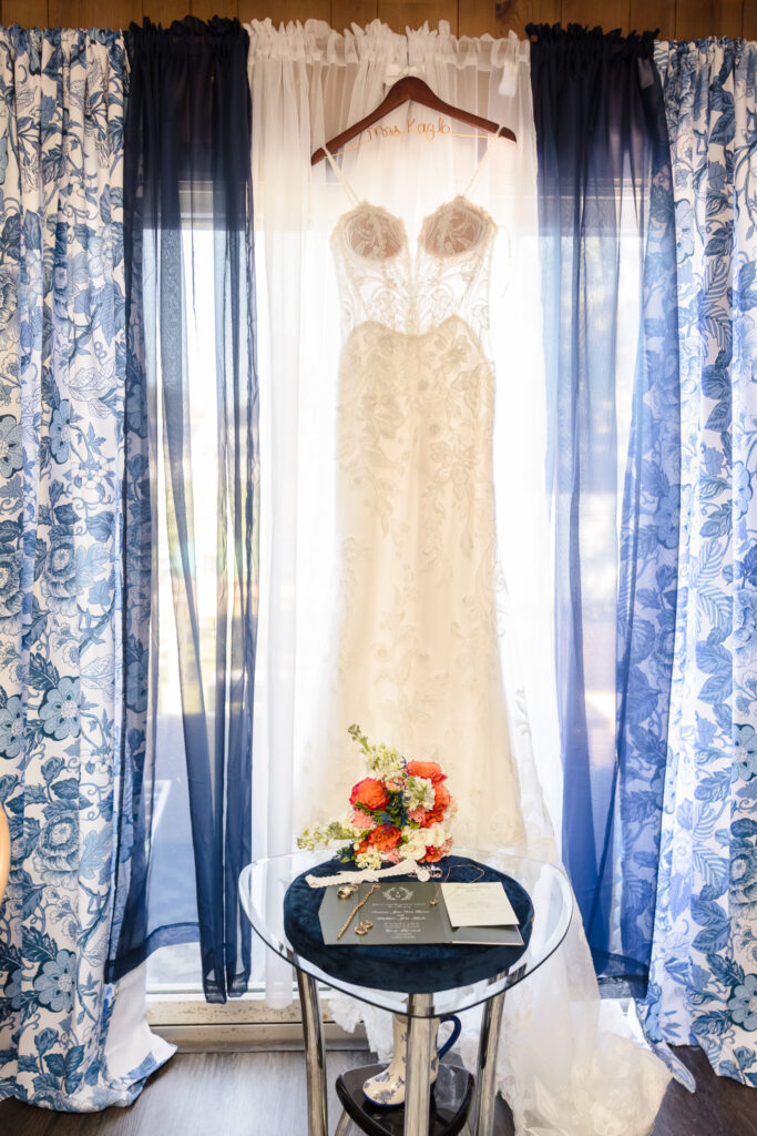 Bridal gown hanging by the window with bouquet, invitation, and jewelry displayed beneath, captured before Sabrina and Matt’s Camp Pa-Qua-Tuck wedding.