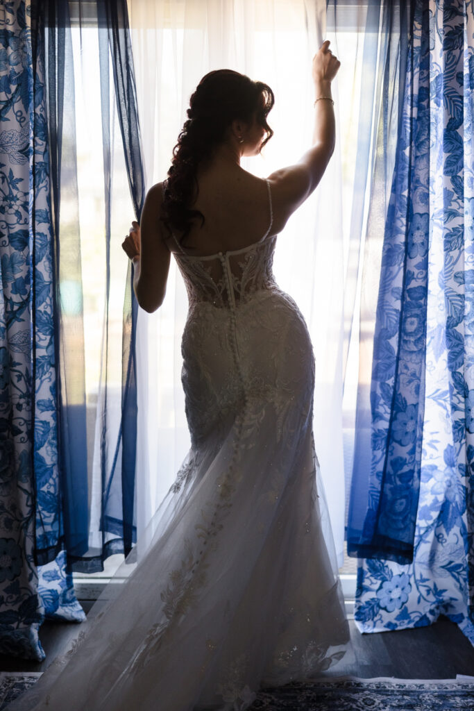 Bride standing in front of window in lace wedding gown, softly lit while getting ready for her Camp Pa-Qua-Tuck wedding.