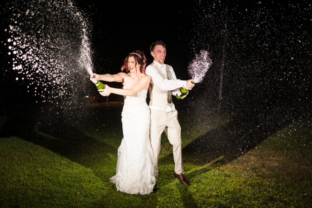 Bride and groom popping champagne at night during their Camp Pa-Qua-Tuck wedding celebration.