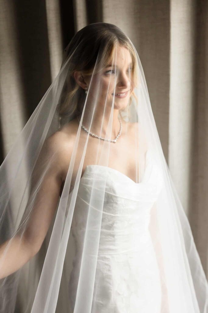 Elegant bride in strapless wedding gown and pearl necklace, softly smiling beneath a sheer veil.