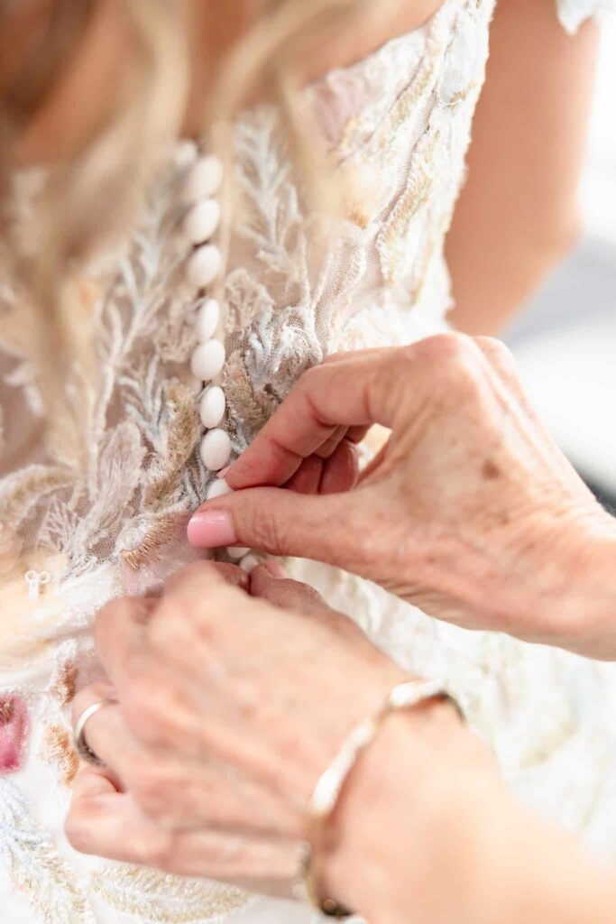 Close-up of hands fastening buttons on a lace wedding gown, capturing intimate bridal preparations.