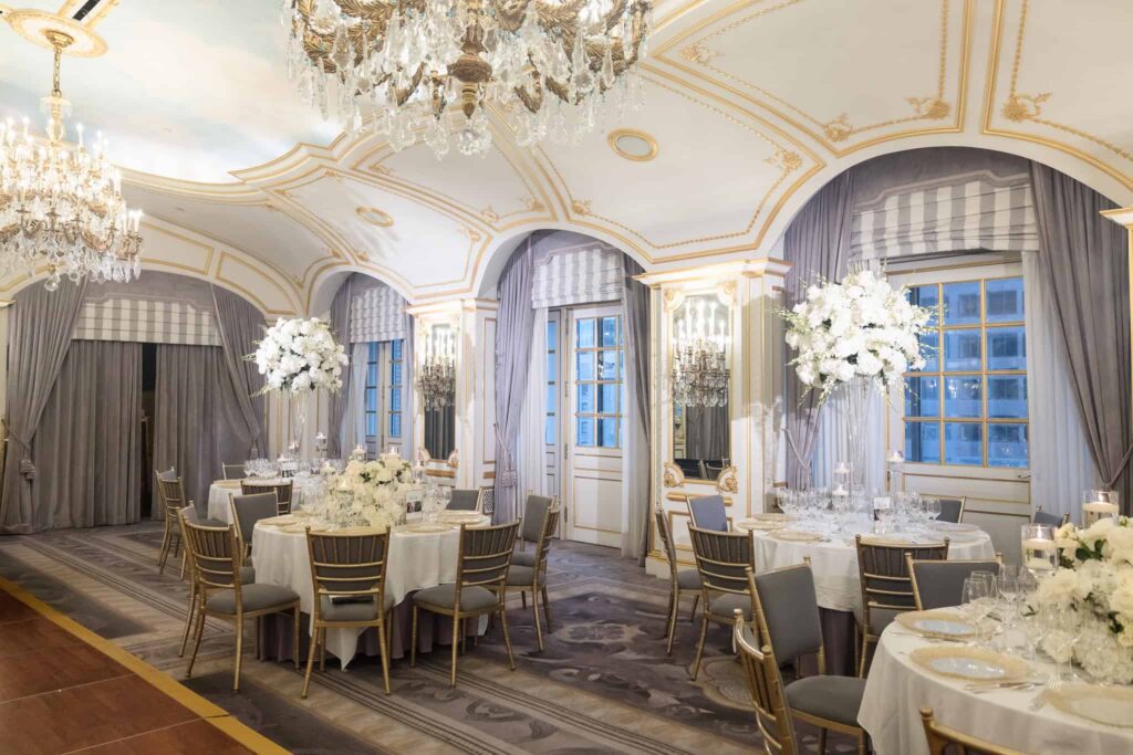 St. Regis New York ballroom set for a wedding reception with tall white floral centerpieces, gold chairs, and ornate crystal chandeliers.