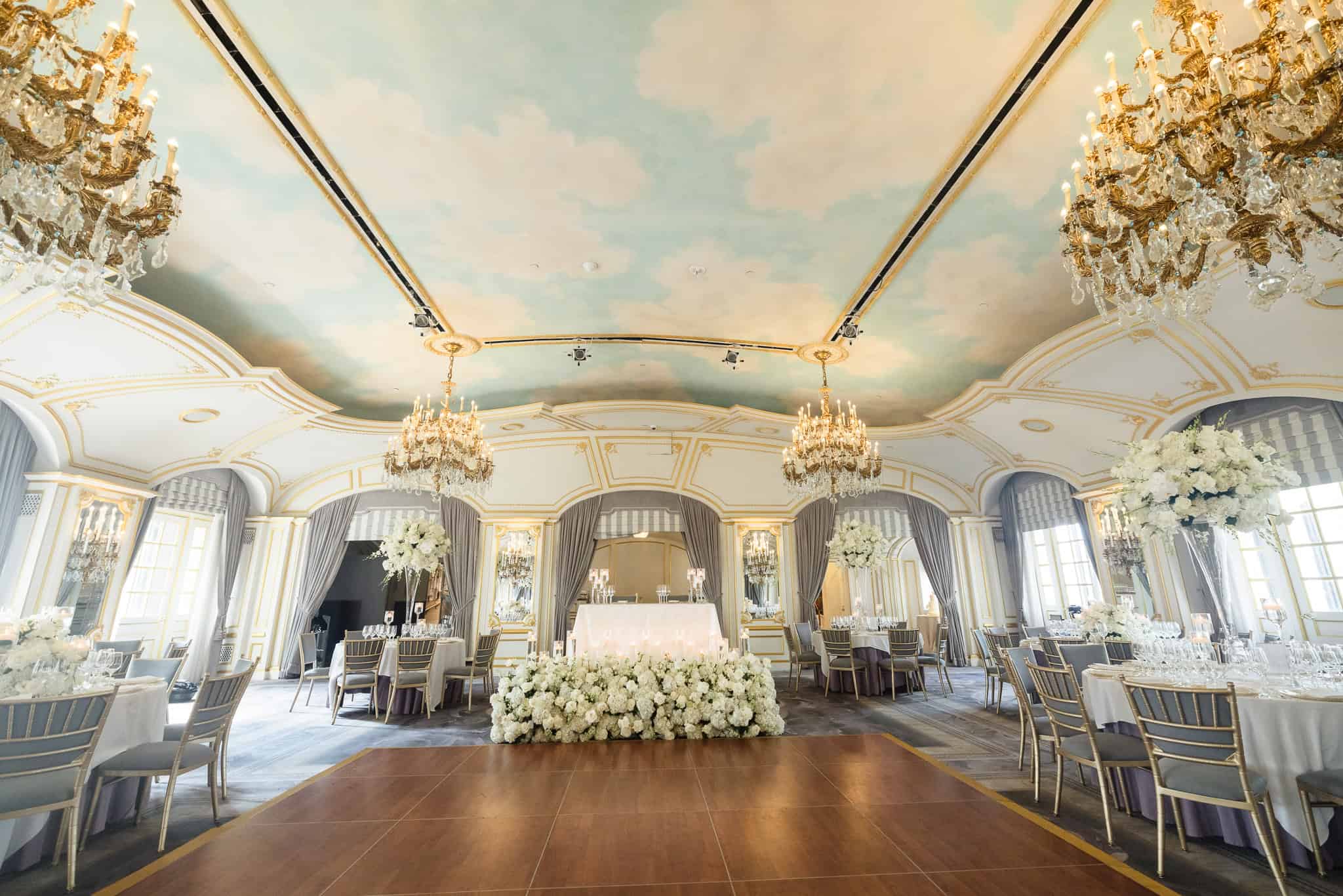 St. Regis New York wedding ballroom with crystal chandeliers, painted sky ceiling, elegant gold chairs, and white floral centerpieces surrounding a romantic sweetheart table.