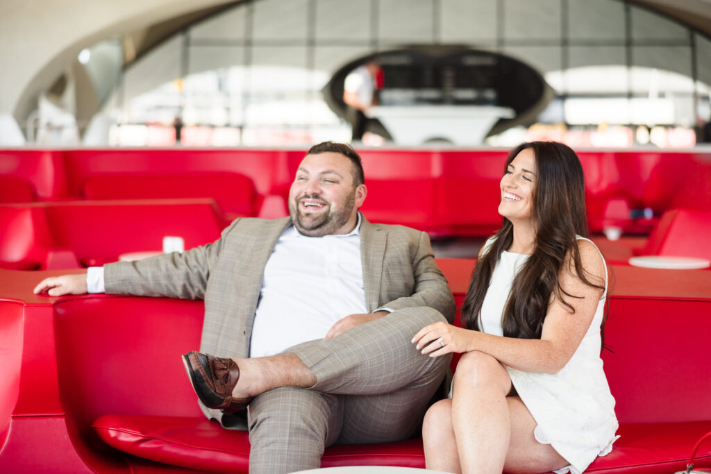 Joyful couple sitting in the iconic red Sunken Lounge at the TWA Hotel during their vintage-inspired engagement session, styled in a tailored suit and white dress for a timeless, mid-century look.
