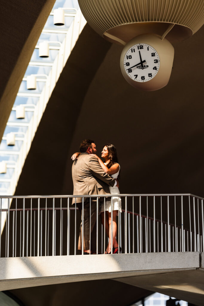 Couple sharing a romantic moment beneath the vintage Vulcain clock at the TWA Hotel, kissed by golden afternoon light inside the iconic mid-century terminal.