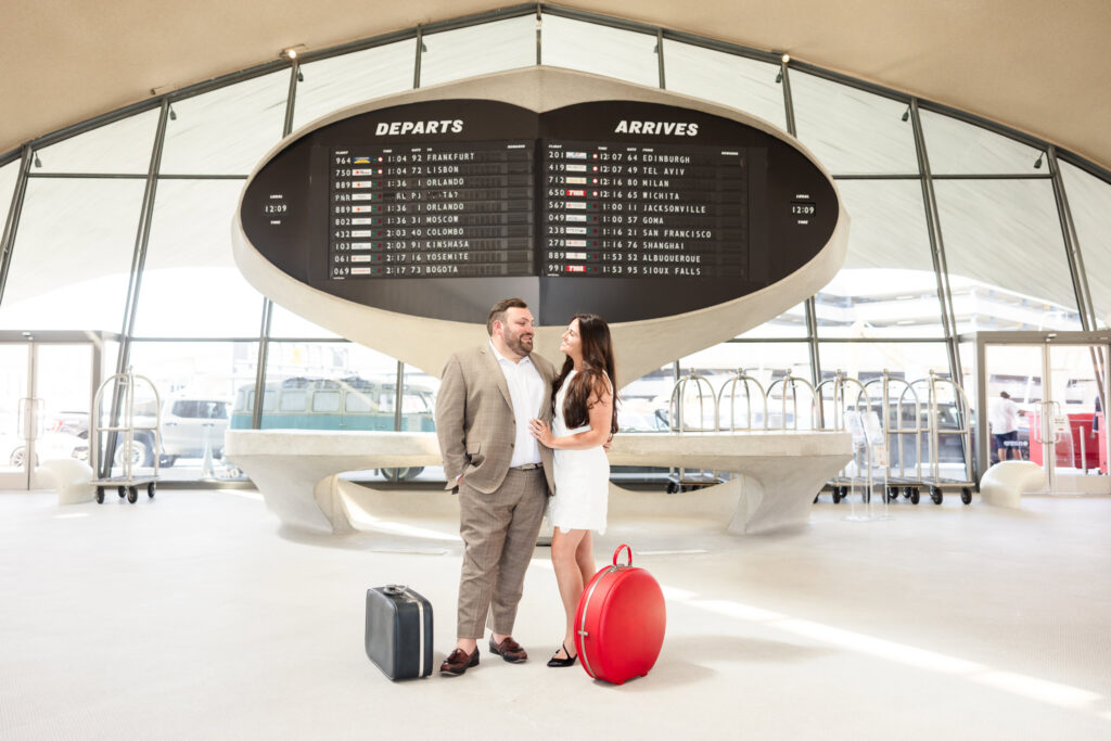 Engaged couple sharing a loving look beneath the retro split-flap departures board at the TWA Hotel, surrounded by vintage suitcases and Jet Age architecture.