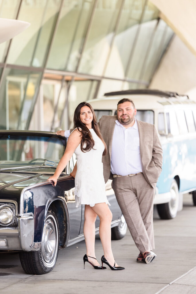 Stylish couple posing beside a vintage black Lincoln Continental at the TWA Hotel’s classic car portico, capturing timeless elegance and retro charm.
