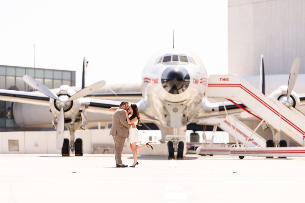 Engaged couple sharing a joyful kiss in front of the vintage TWA Lockheed Constellation airplane, with the bride playfully lifting one foot in a classic romantic pose on the hotel tarmac.