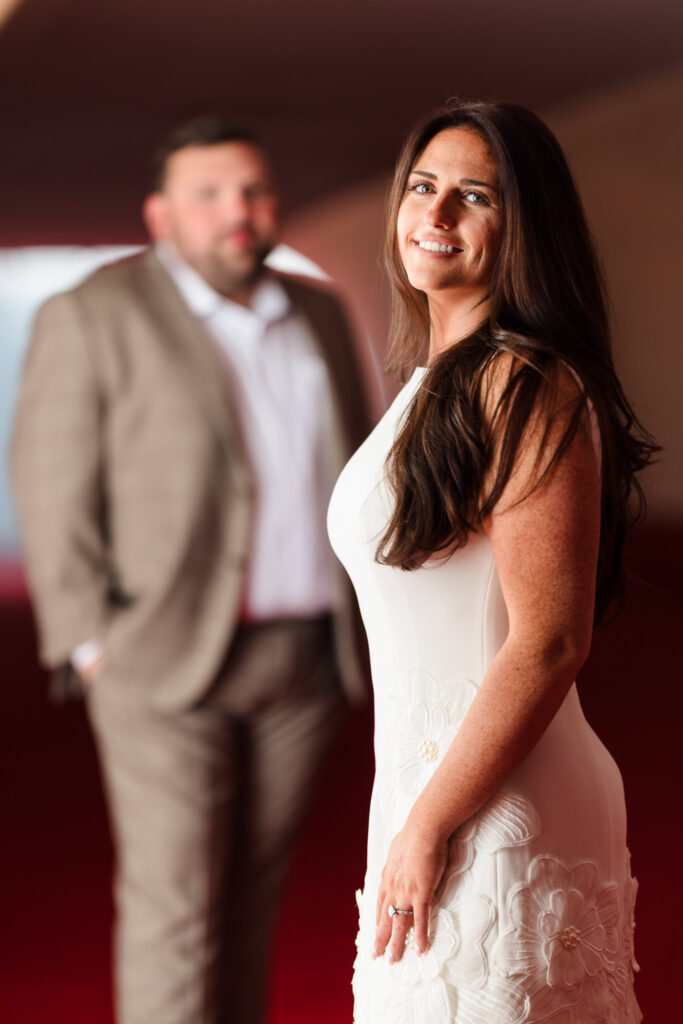 Bride-to-be smiling softly in the foreground, wearing a floral-detailed white dress, with her fiancé blurred in the background at the TWA Hotel’s red-carpeted tunnel.