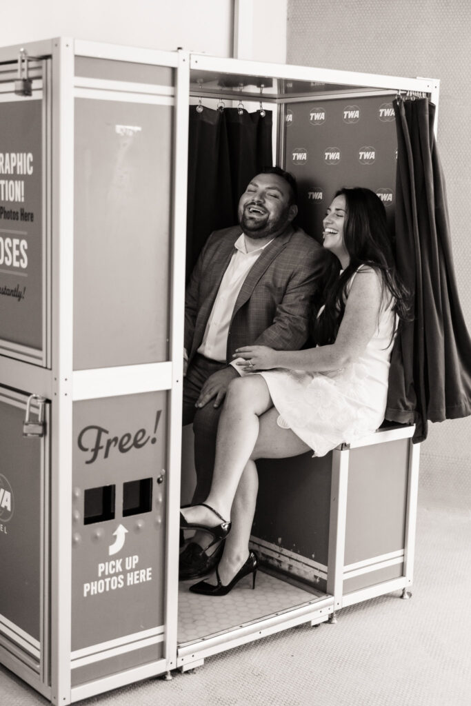Playful black-and-white moment of an engaged couple laughing together inside a vintage-style photo booth at the TWA Hotel, capturing candid joy and retro charm.