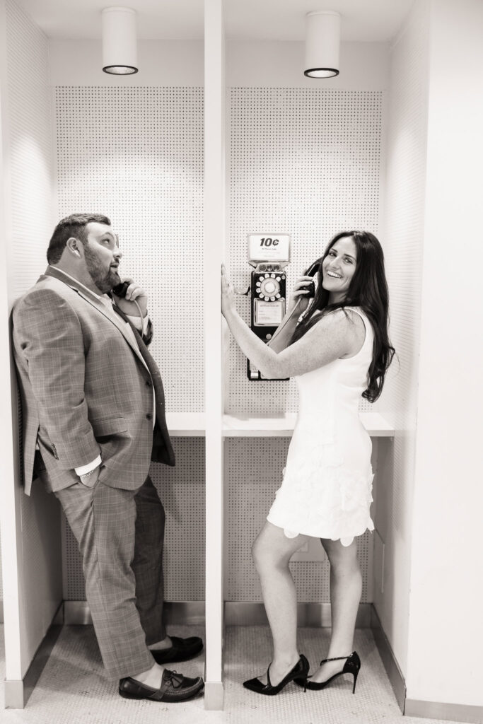 Whimsical black-and-white engagement portrait of a couple playfully posing in vintage phone booths at the TWA Hotel, channeling retro romance and mid-century nostalgia.