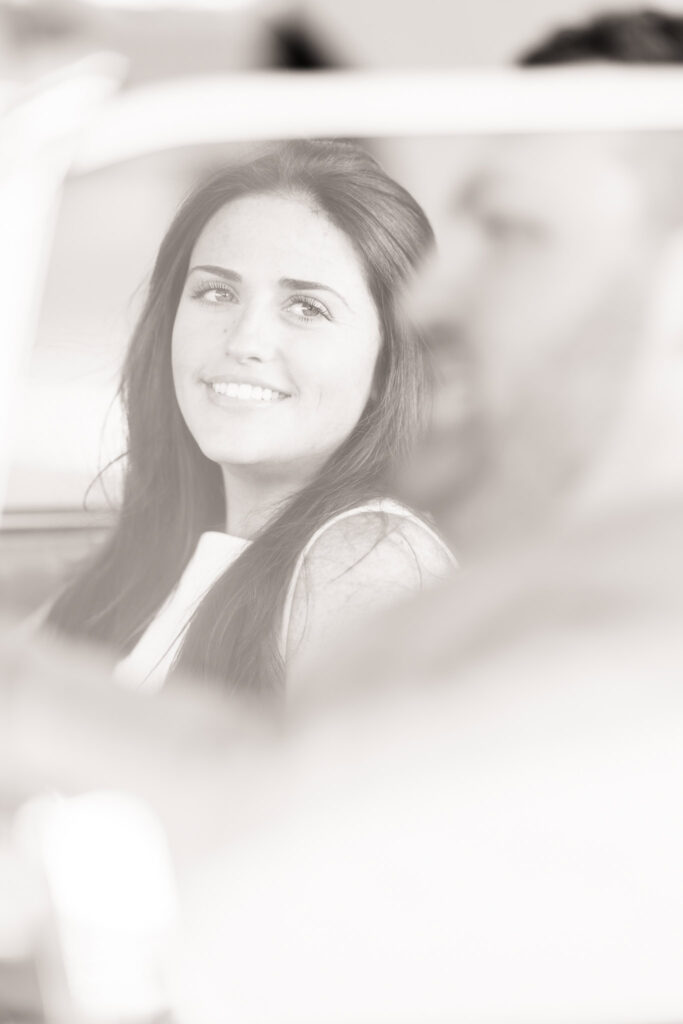 Soft-focus black-and-white portrait of a bride-to-be smiling through a vintage car window, capturing a quiet, intimate moment during her TWA Hotel engagement session.