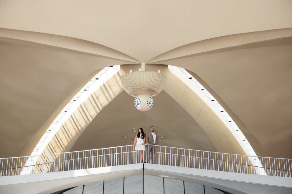 Engaged couple standing on the mezzanine of the TWA Hotel beneath its iconic mid-century clock and sweeping architectural arches, bathed in soft afternoon light.