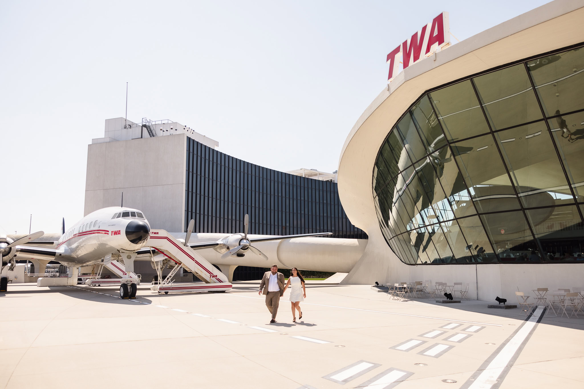Engaged couple walking hand-in-hand on the tarmac outside the TWA Hotel, framed by the vintage Connie aircraft and the hotel’s iconic mid-century modern architecture.