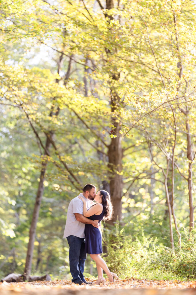 Couple kissing in the woods while standing on fallen leaves. 