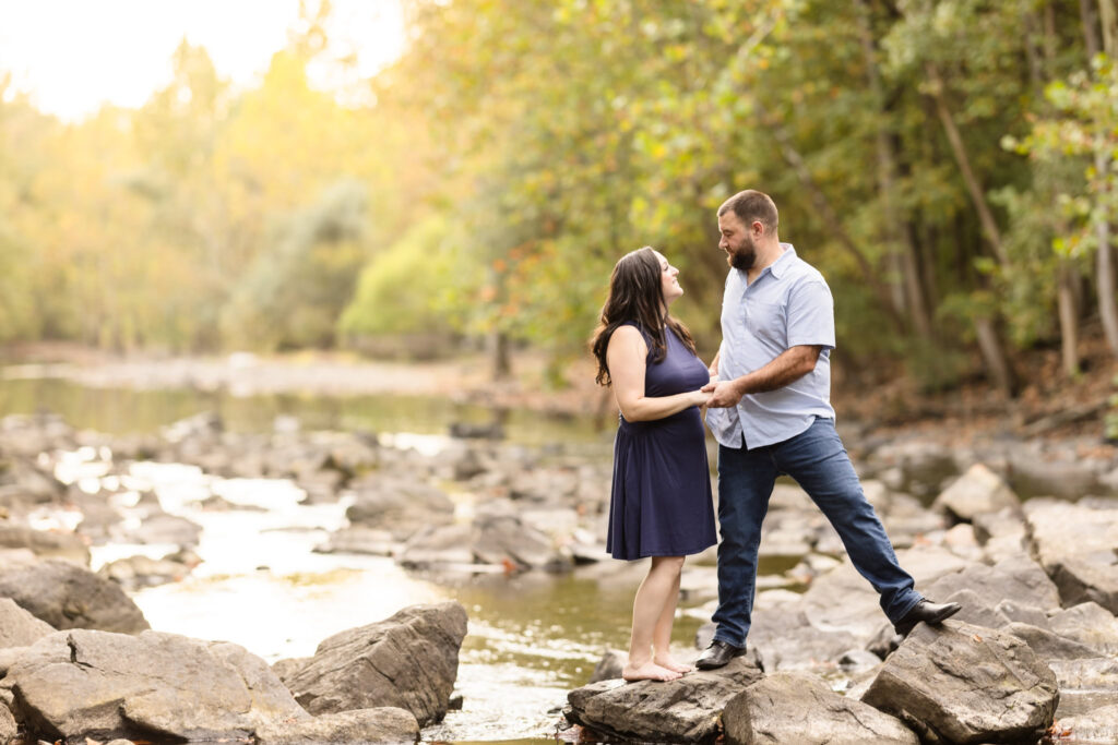 Couple standing on rocks in stream at a fall engagement session with changing leaves and a sun flare behind them. 