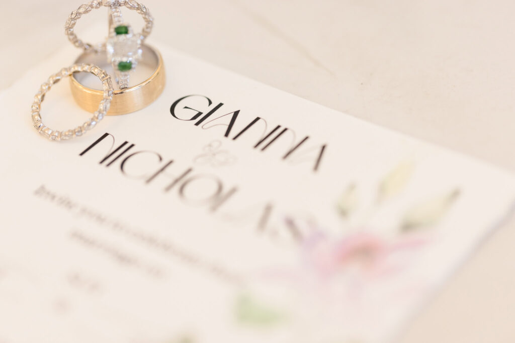 Close-up of wedding rings, including an emerald-accented engagement ring, resting on a floral invitation card featuring the names of Bride and the Groom.