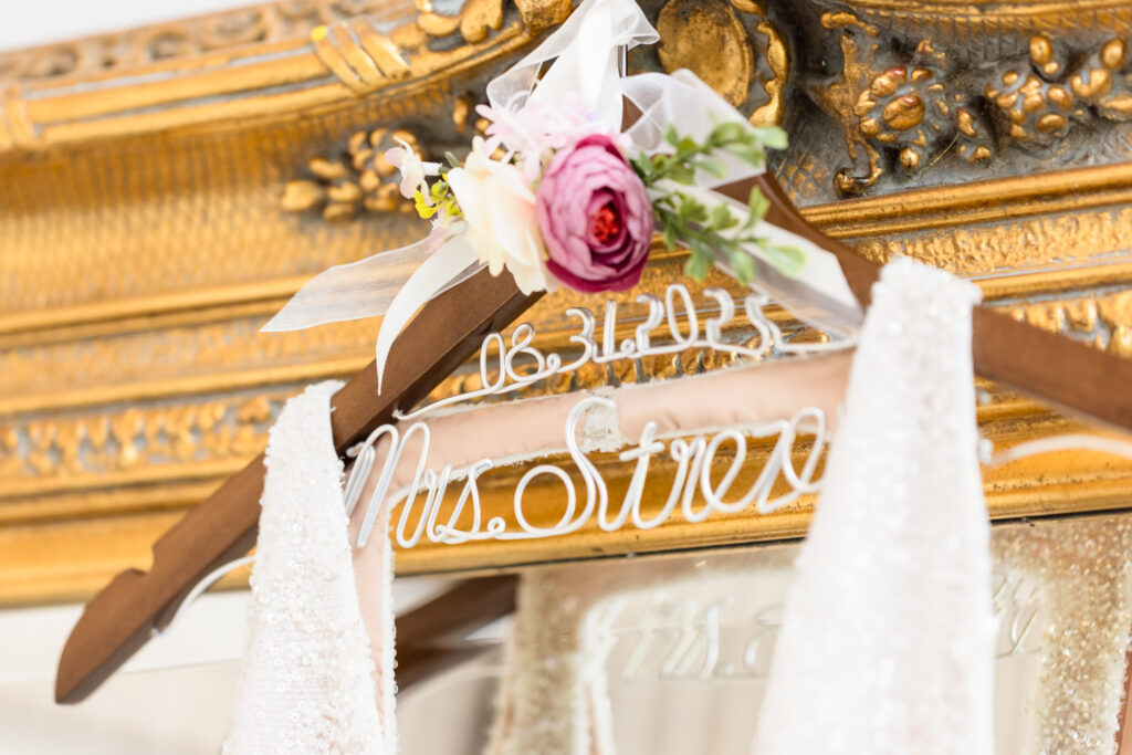 Close-up of a personalized wedding dress hanger reading “Mrs. Strez” and the date “06.30.2023,” adorned with a ribbon and floral accent, holding a sparkly bridal gown in front of an ornate gold mirror.